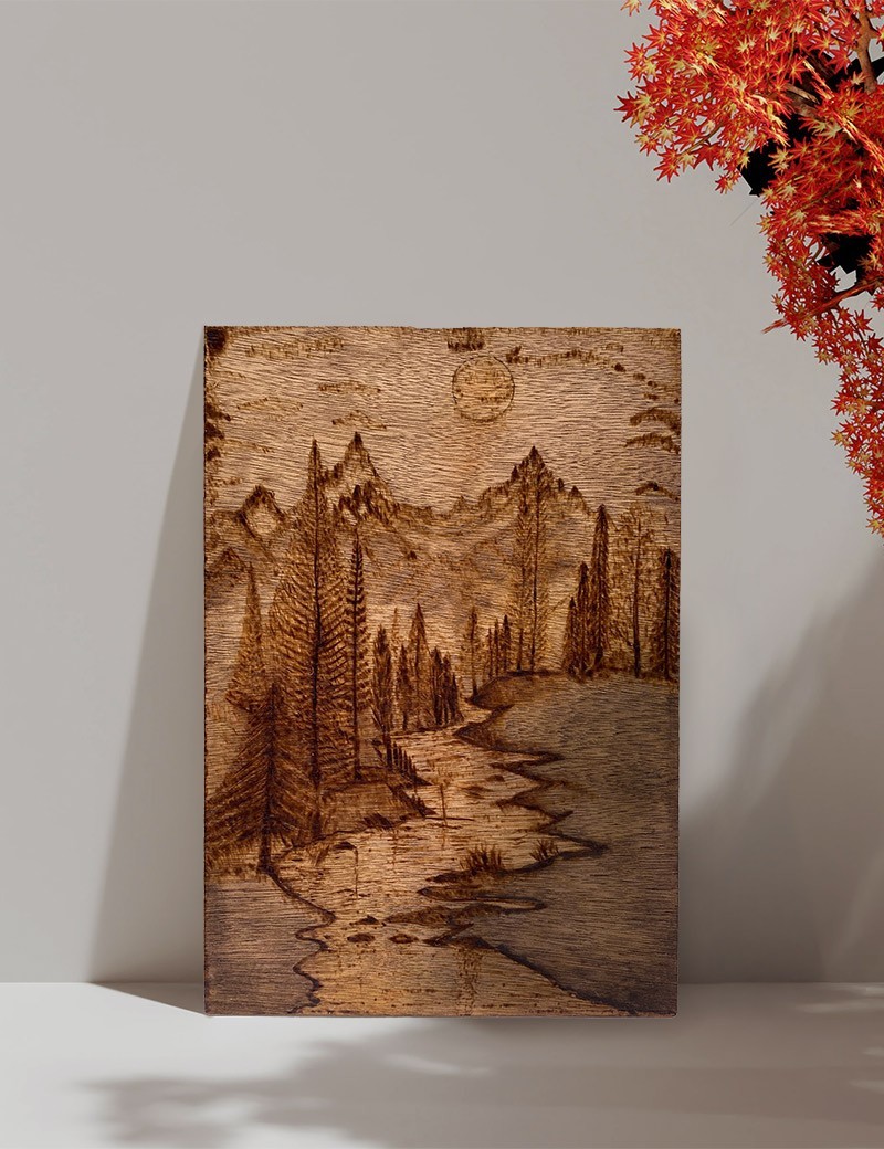 A4 woodburning art of a mountain landscape leaning against a white wall with autumn leaves.