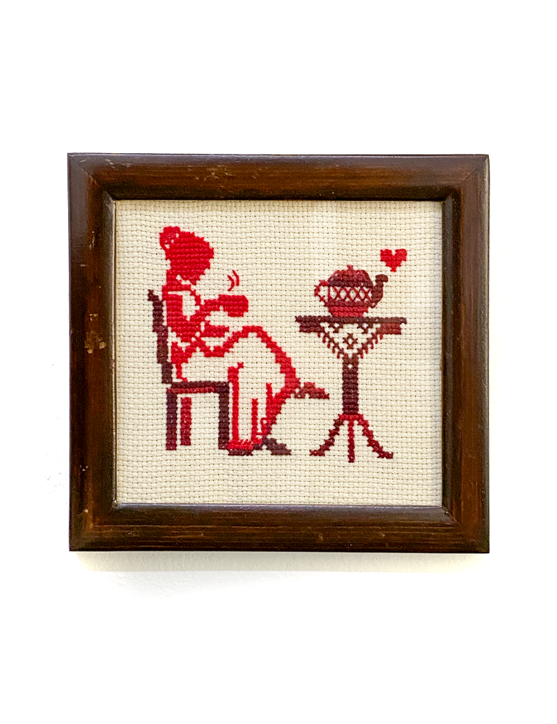 Front view of framed red cross-stitch of a woman drinking tea.