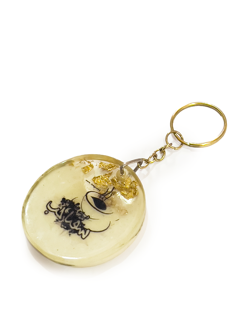 Round cream-colored resin keychain featuring black Arabic calligraphy and gold foil accents.