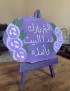 Purple painted rustic wooden plaque with white floral design and Arabic home blessing calligraphy.