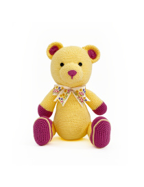 Full front view of a sitting yellow and purple crochet teddy bear with a floral bow.