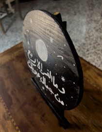 Black wooden round painted with moon, stars, and white Arabic script about beauty and eyes.
